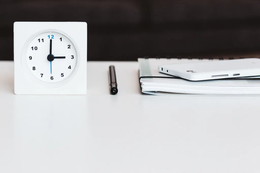Small desk clock beside a pen, planner and phone on a white desk