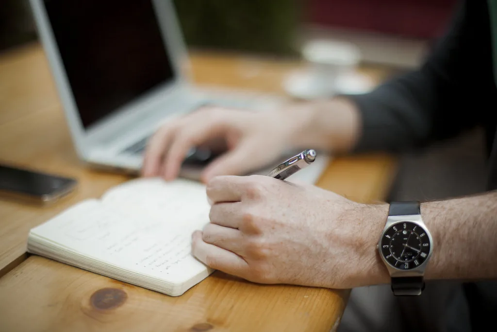 A person writing in a notebook beside a laptop at a wooden desk