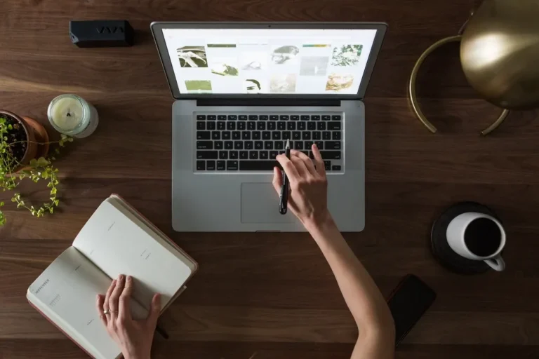 Hands working at a laptop beside a notebook and coffee, seen from above