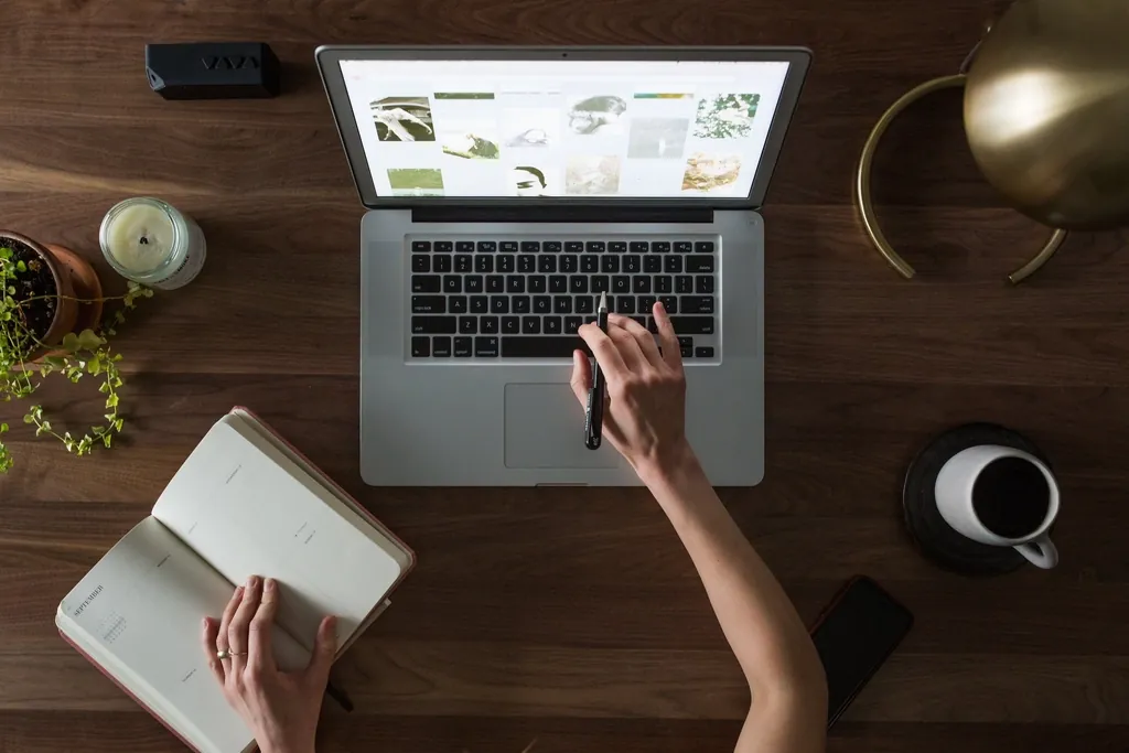 Hands working at a laptop beside a notebook and coffee, seen from above